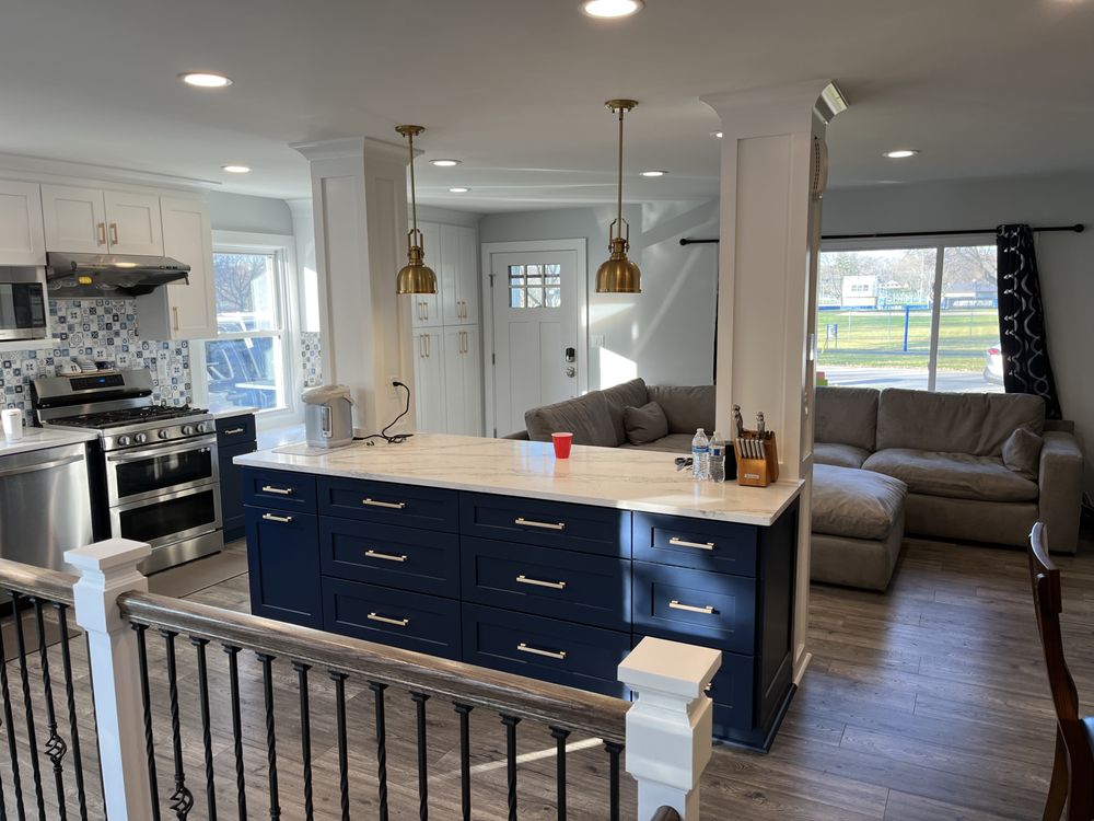 Modern kitchen and living area with open layout. Features a navy island with marble top, brass pendant lights, white cabinetry, and cozy gray couches. Bright, inviting atmosphere.