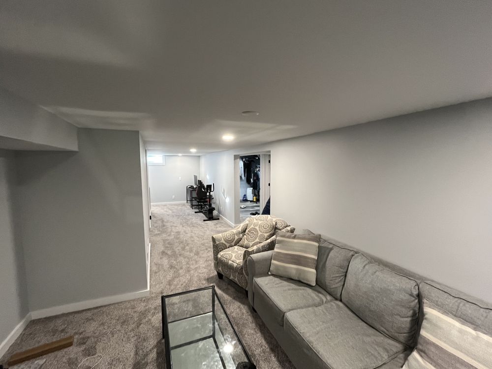 Basement room with gray carpet, featuring gray sofas with striped cushions, a glass coffee table, and a distant view of exercise equipment.