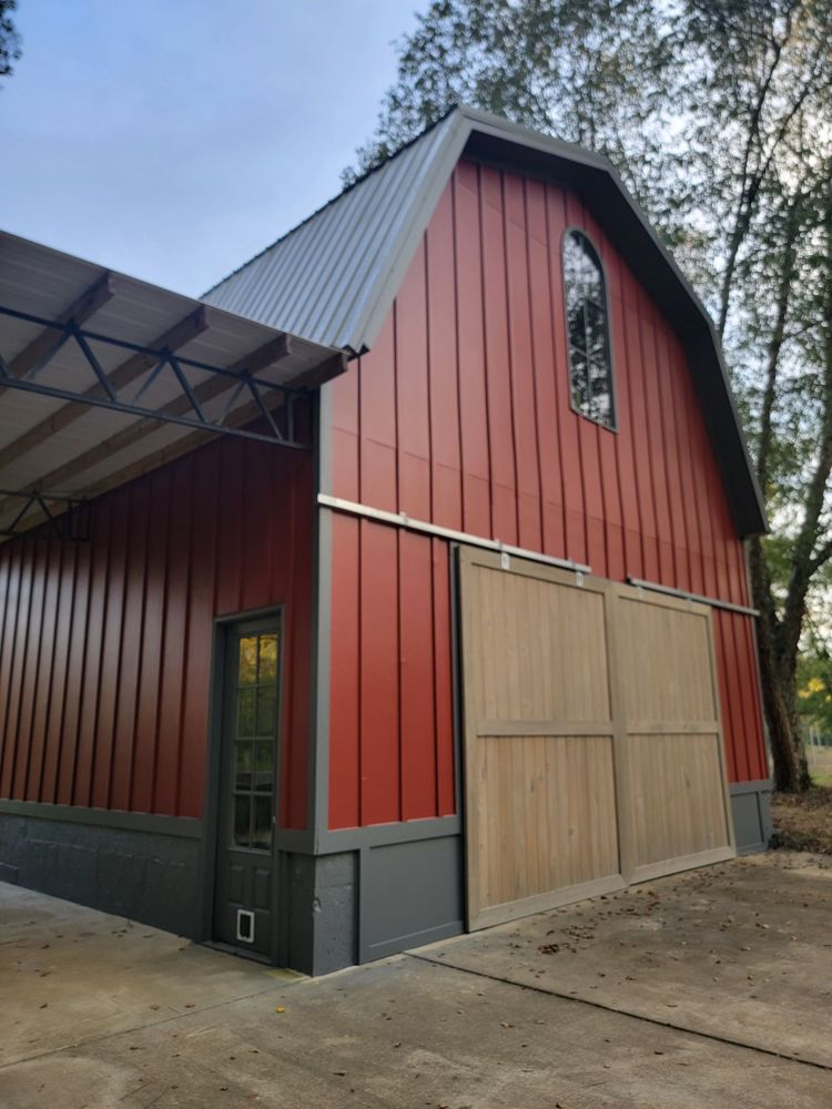 A red barn with vertical paneling and a metal roof stands on a concrete slab. The barn has large wooden sliding doors and a small black-framed window. Trees surround the structure, adding a rustic, serene feel.