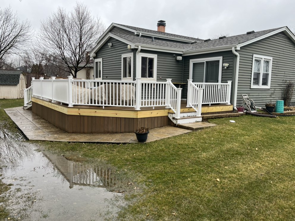 Gray house with a white-railed deck, situated in a grassy yard. A cloudy sky looms overhead, with wet patches and puddles on the ground.
