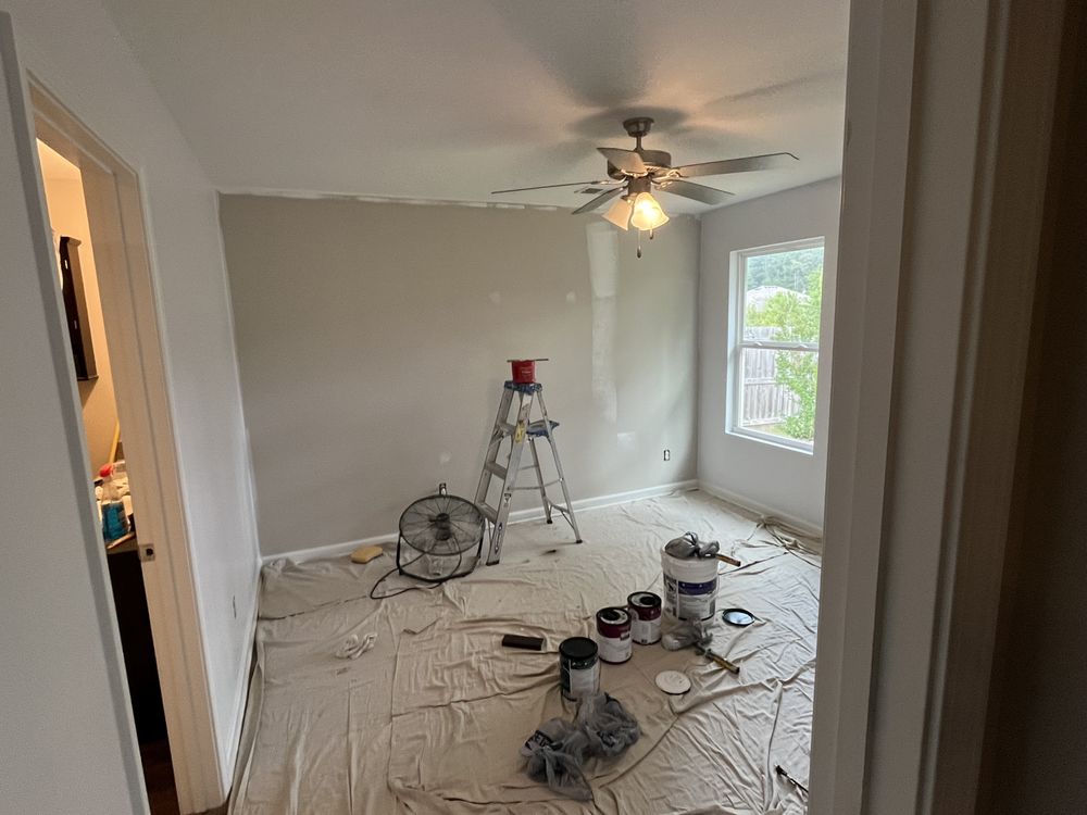A room under renovation with a drop cloth on the floor, a ladder, paint cans, and a brush. Walls are partially painted beige. A ceiling fan is overhead.