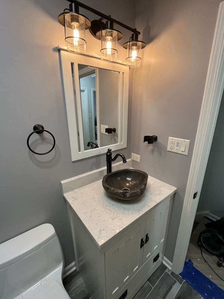 Modern bathroom with a white vanity, marble countertop, and a black vessel sink. Above is a mirror with three industrial-style light fixtures.